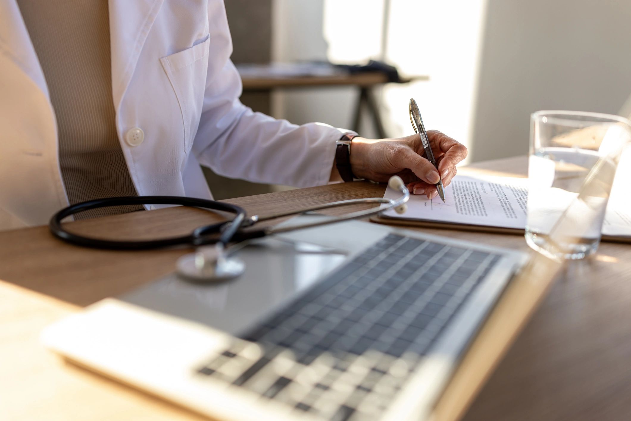 Doctor signing medical documents at a desk with a laptop and stethoscope in a bright healthcare office